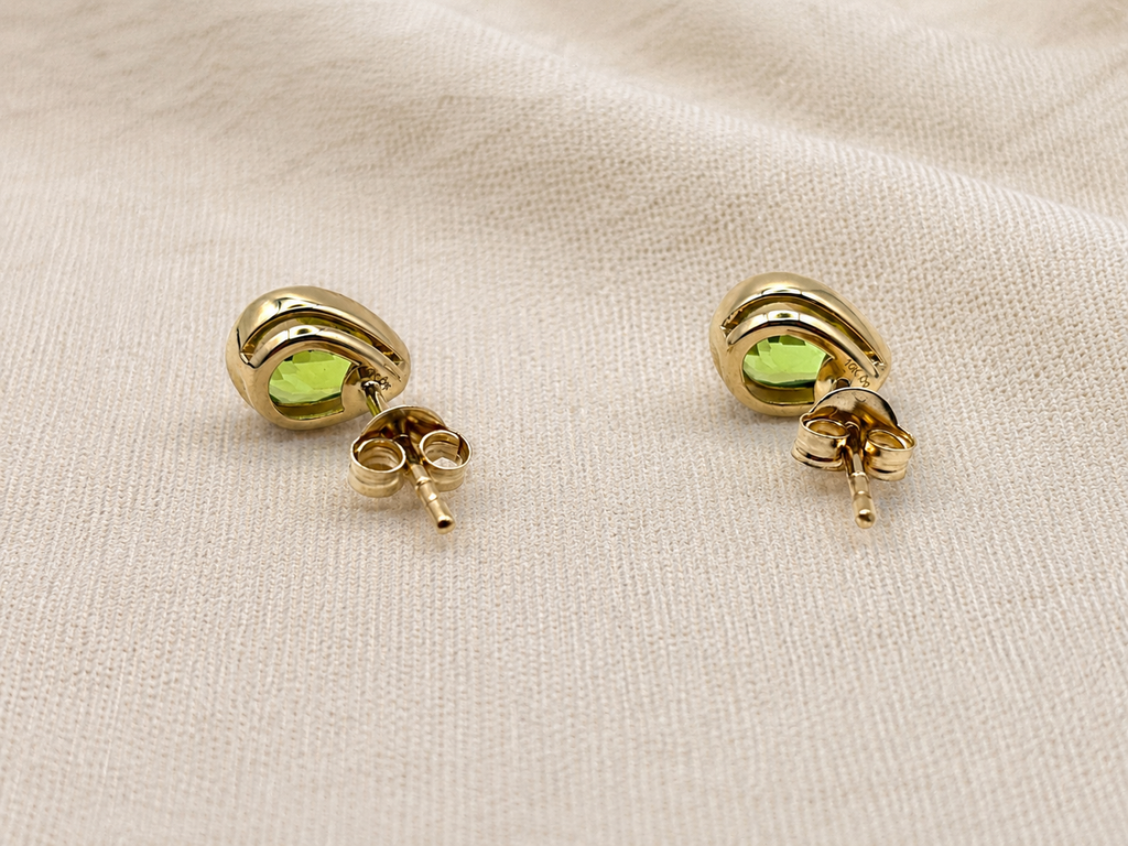 A pair of solid 10k yellow gold stud earrings with oval natural green peridot gemstones by Ophelisse, displayed on a textured beige fabric background, with the back showing Ophelisse trademarks.