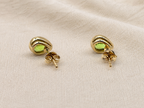 A pair of solid 10k yellow gold stud earrings with oval natural green peridot gemstones by Ophelisse, displayed on a textured beige fabric background, with the back showing Ophelisse trademarks.