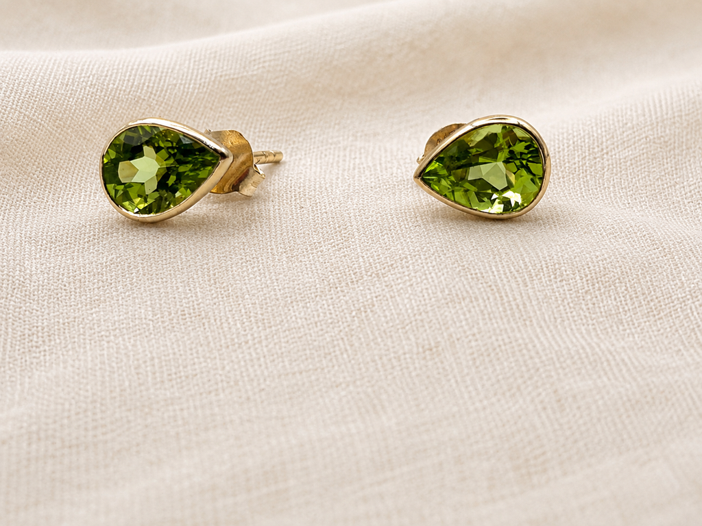 A pair of 10k solid yellow gold teardrop stud earrings with faceted natural green peridot gemstones by Ophelisse, resting on a textured beige fabric background.