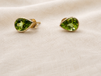 A pair of 10k solid yellow gold teardrop stud earrings with faceted natural green peridot gemstones by Ophelisse, resting on a textured beige fabric background.