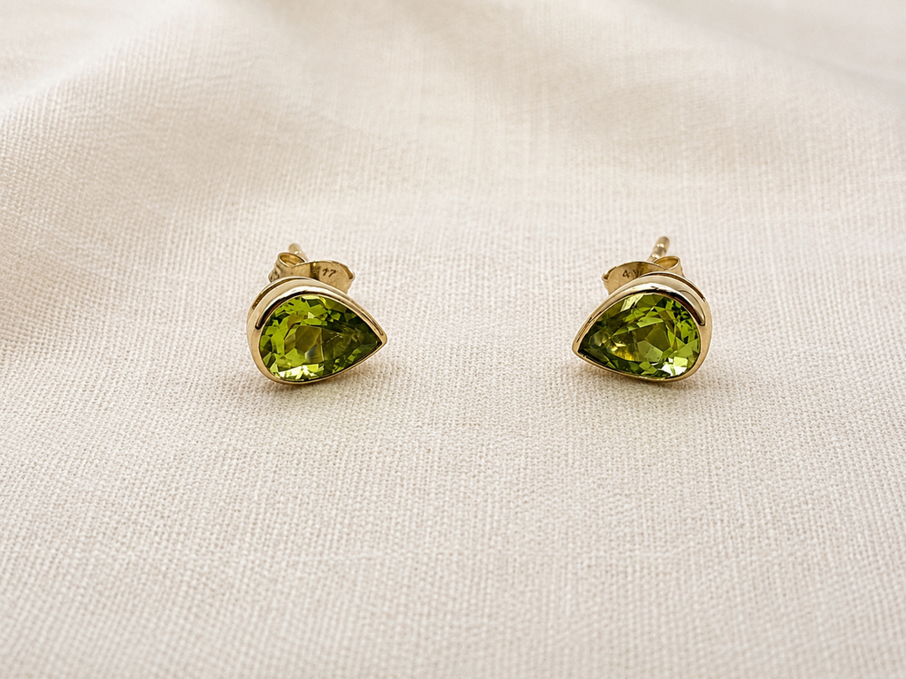 A pair of solid 10k yellow gold stud earrings with teardrop-shaped natural green peridot gemstones by Ophelisse, displayed on a textured beige fabric background.