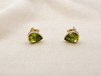 A pair of solid 10k yellow gold stud earrings with teardrop-shaped natural green peridot gemstones by Ophelisse, displayed on a textured beige fabric background.