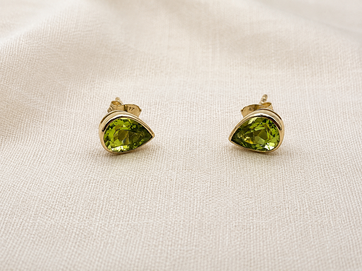 A pair of solid 10k yellow gold stud earrings with teardrop-shaped natural green peridot gemstones by Ophelisse, displayed on a textured beige fabric background.
