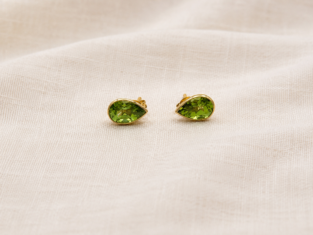 A pair of luxurious 10k solid yellow gold stud earrings with marquise-cut natural green peridot gemstones by Ophelisse, resting on a textured beige linen fabric background.
