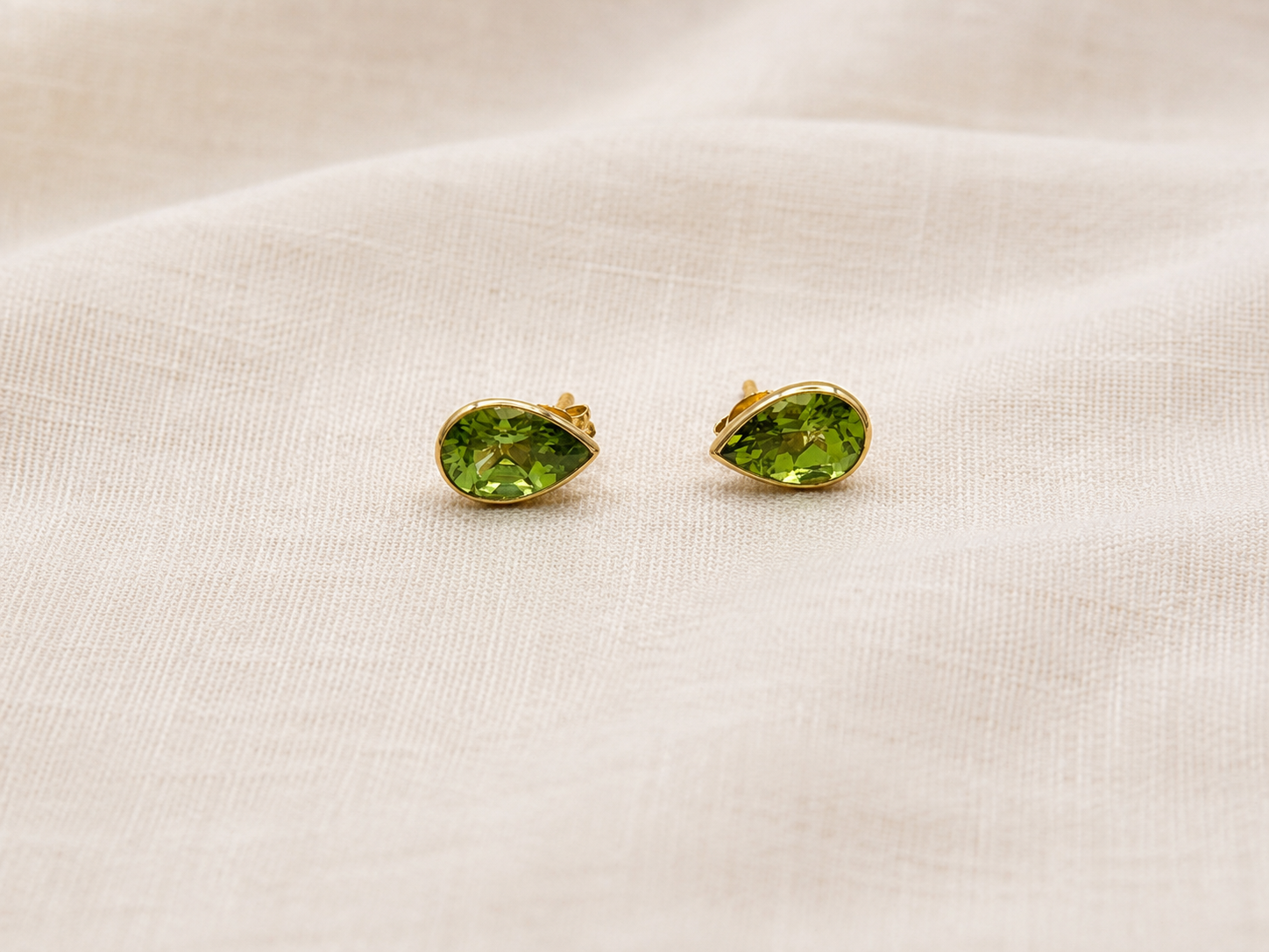 A pair of luxurious 10k solid yellow gold stud earrings with marquise-cut natural green peridot gemstones by Ophelisse, resting on a textured beige linen fabric background.