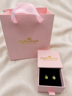 A pair of small natural green teardrop gemstones earrings displayed in an open pink jewelry box with a pink ribbon pull, next to a larger pink Ophelisse shopping bag on a light fabric background.