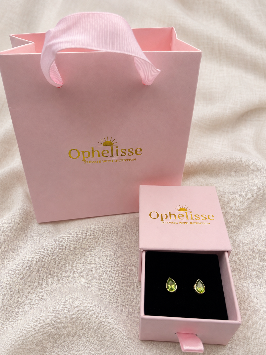 A pair of small natural green teardrop gemstones earrings displayed in an open pink jewelry box with a pink ribbon pull, next to a larger pink Ophelisse shopping bag on a light fabric background.