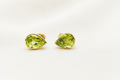 A pair of 10k solid yellow gold stud earrings with a luxurious faceted green natural peridot gemstones by Ophelisse, displayed on a textured beige fabric background.