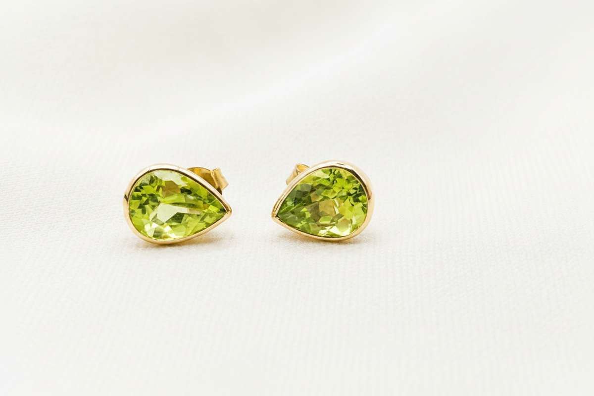 A pair of 10k solid yellow gold stud earrings with a luxurious faceted green natural peridot gemstones by Ophelisse, displayed on a textured beige fabric background.