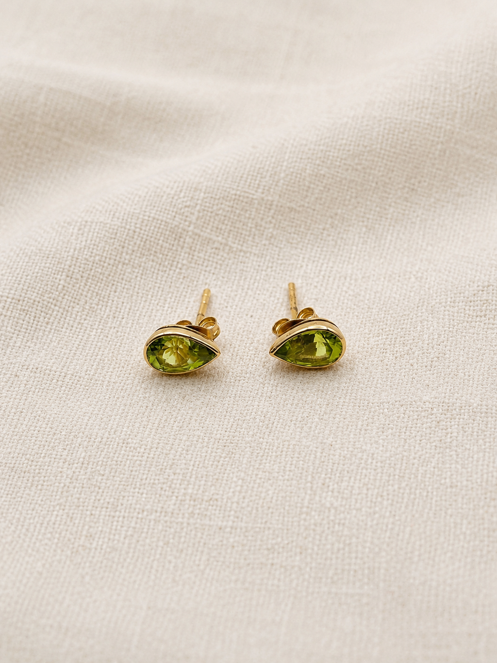 A pair of solid, elegant 10k yellow gold stud earrings with marquise-cut natural green Paridot gemstones by Ophelisse, displayed on a textured beige linen fabric background.