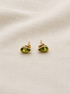 A pair of solid, elegant 10k yellow gold stud earrings with marquise-cut natural green Paridot gemstones by Ophelisse, displayed on a textured beige linen fabric background.