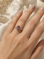 A close-up of a hand wearing a delicate solid 10k yellow gold ring with a small oval purple amethyst gemstone by Ophelisse, set against a soft beige textured fabric background.