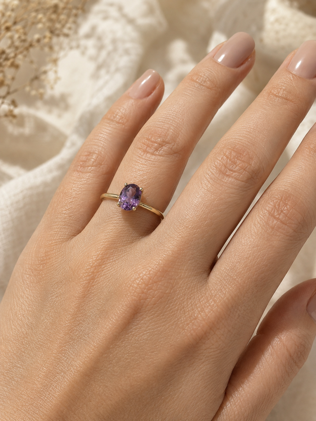 A close-up of a hand wearing a delicate solid 10k yellow gold ring with a small oval purple amethyst gemstone by Ophelisse, set against a soft beige textured fabric background.