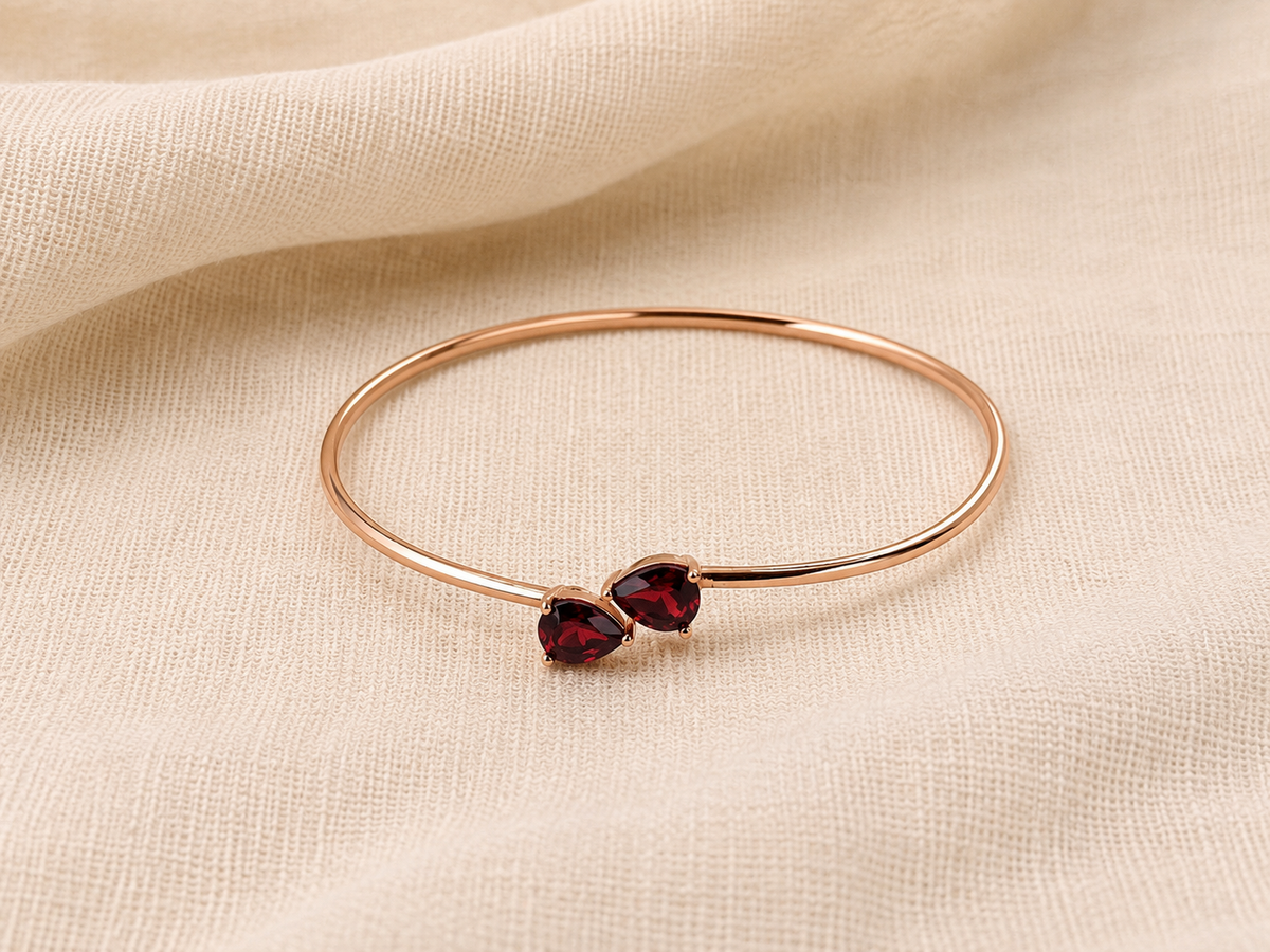 A solid 10k rose gold bangle bracelet with two small heart-shaped dark red natural garnet gemstones by Ophelisse, displayed flat on a textured beige fabric background showing the top view of the bangle and natural garnet.