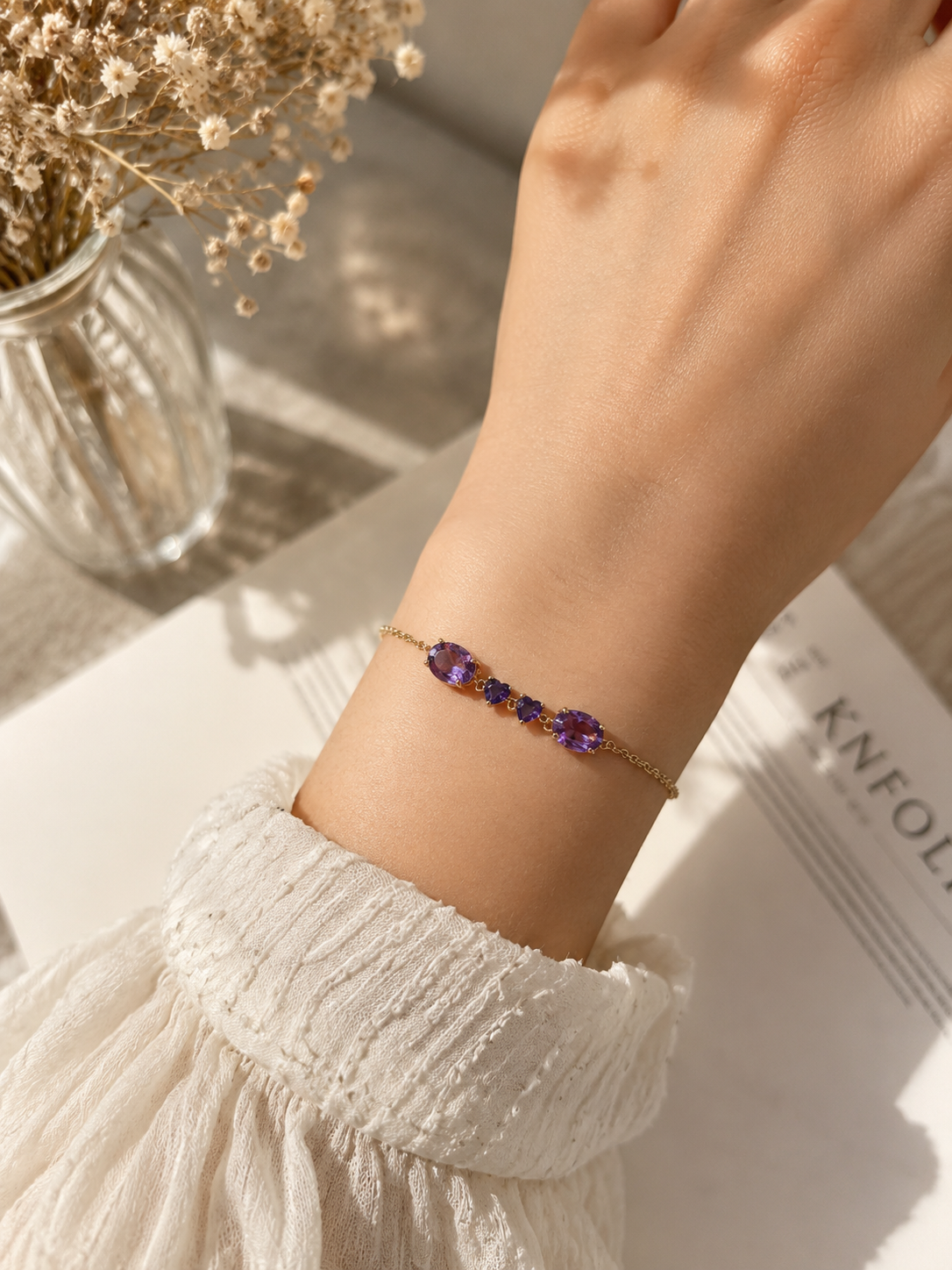 A solid 10k yellow gold bracelet by Ophelisse with two oval-shaped and two heart-shaped natural purple amethyst gemstones worn on a person's wrist, styled with a white textured dress and dried flowers in a glass vase.