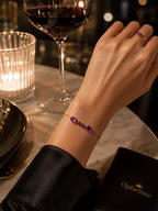 A close-up lifestyle shot of a woman's wrist wearing a solid 10k gold bracelet with two oval and two heart natural  amethyst  gemstones. The scene is set on a marble table with a glass of red wine, a lit candle, and an Ophelisse brand box nearby.