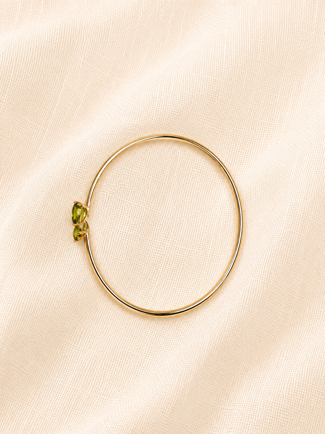 A solid 10k yellow gold bangle bracelet with two natural green gemstone by Ophelisse, displayed on a textured cream-colored fabric background, showing side view