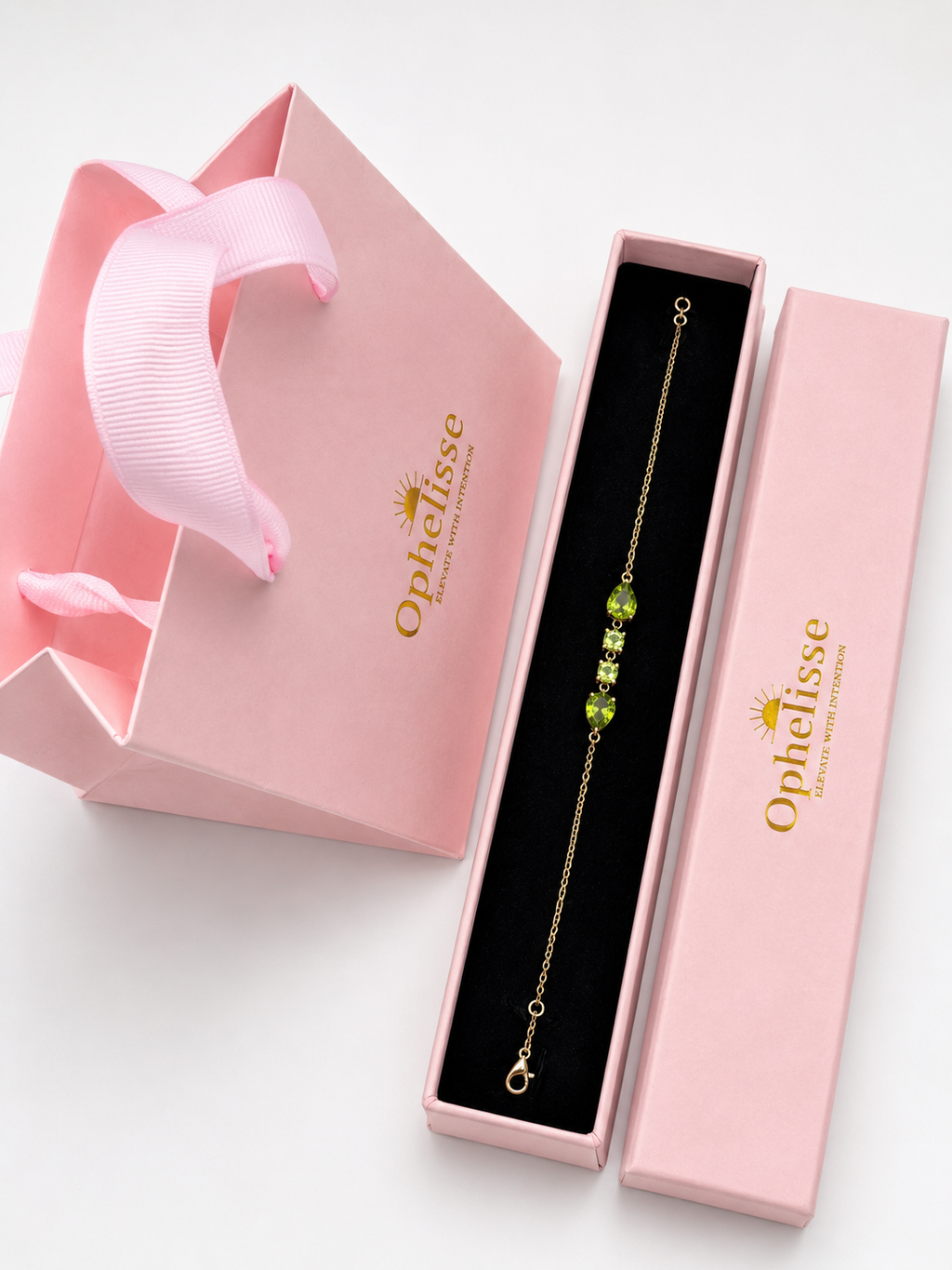 A solid 10k gold chain bracelet with natural green peridot gemstone accents, presented in an open pink jewelry box next to its closed lid and a pink gift bag with the Ophelisse logo.