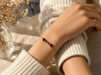 A delicate solid 10k rose gold chain bracelet with two heart-shaped and two round-shaped natural red garnet gemstone worn on a person's wrist, styled with a cream ribbed knit sweater and dried flowers in a glass vase.