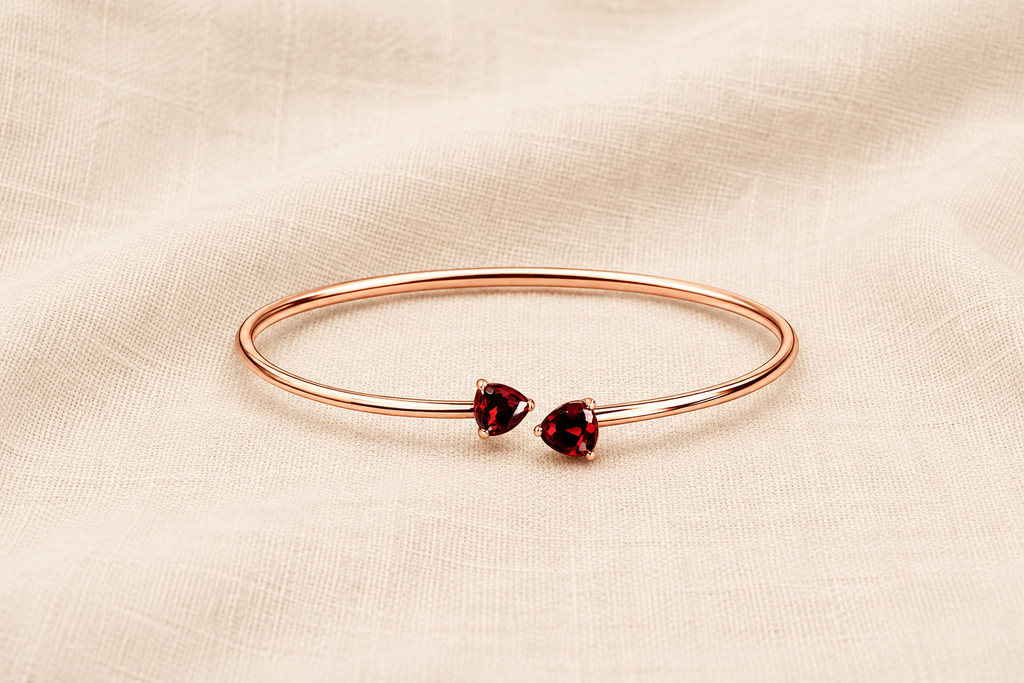 A solid 10k rose gold bangle bracelet with two small heart-shaped natural dark red garnet gemstones by Ophelisse, displayed flat on a textured beige fabric background showing the top view of the natural bangle and natural garnet stones