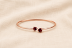 A solid 10k rose gold bangle bracelet with two small heart-shaped natural dark red garnet gemstones by Ophelisse, displayed flat on a textured beige fabric background showing the top view of the natural bangle and natural garnet stones