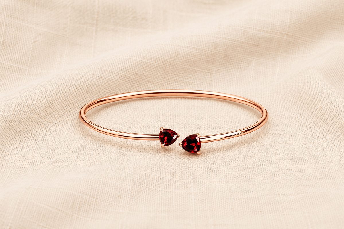 A solid 10k rose gold bangle bracelet with two small heart-shaped natural dark red garnet gemstones by Ophelisse, displayed flat on a textured beige fabric background showing the top view of the natural bangle and natural garnet stones