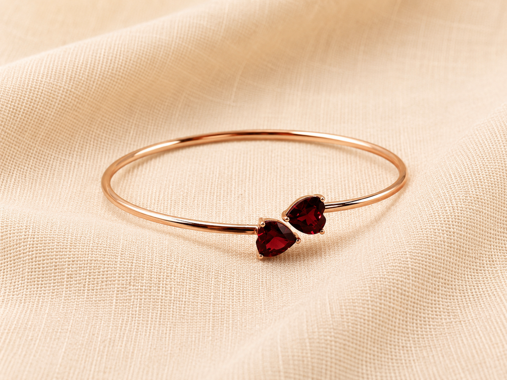 A solid 10k rose gold bangle bracelet with two small heart-shaped dark red natural garnet gemstones by Ophelisse, displayed flat on a textured beige fabric background showing the top view of the  bangle and natural garnet.