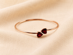 A solid 10k rose gold bangle bracelet with two small heart-shaped dark red natural garnet gemstones by Ophelisse, displayed flat on a textured beige fabric background showing the top view of the  bangle and natural garnet.