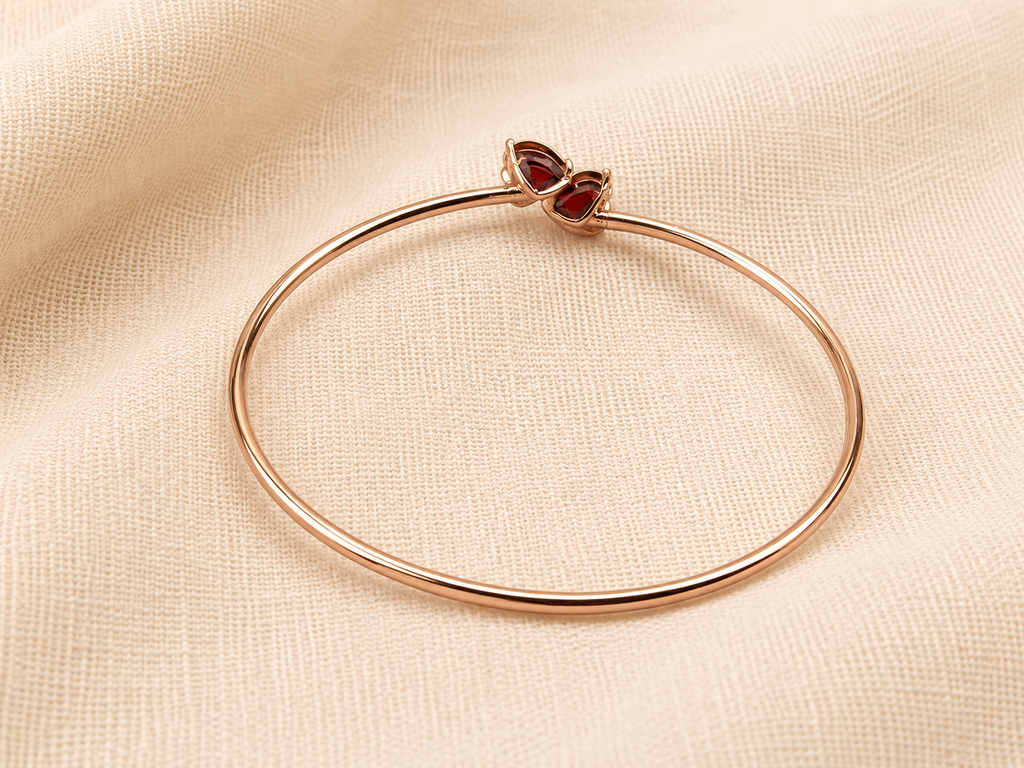 A solid 10k rose gold bangle bracelet with a heart-shaped natural red garnet gemstone clasp by Ophelisse, displayed flat on a textured beige fabric background showing the bottom of the natural garnet stones