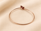 A solid 10k rose gold bangle bracelet with a heart-shaped natural red garnet gemstone clasp by Ophelisse, displayed flat on a textured beige fabric background showing the bottom of the natural garnet stones