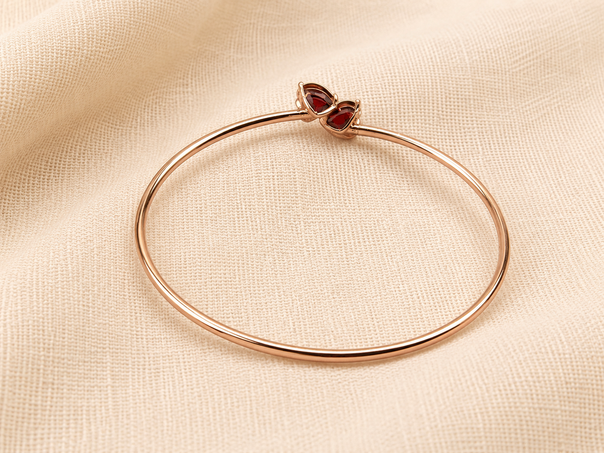 A solid 10k rose gold bangle bracelet with a heart-shaped natural red garnet gemstone clasp by Ophelisse, displayed flat on a textured beige fabric background showing the bottom of the natural garnet stones