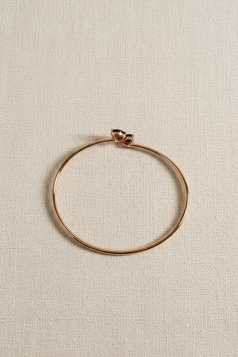 Solid 10k rose gold bangle by Ophelisse, featured two natural red garnet gemstones, flat top view on a light beige linen background