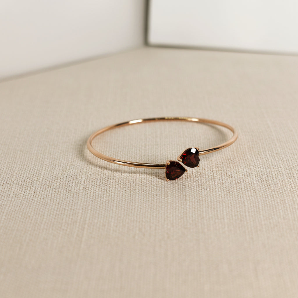 Top flat view of Ophelisse solid 10k rose gold bangle with two heart-shaped natural garnet gemstones on a beige linen surface