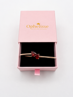 A solid 10k rose gold bangle bracelet with two red heart-shaped natural garnet gemstones, presented in an open pink Ophelisse gift box on a white background.
