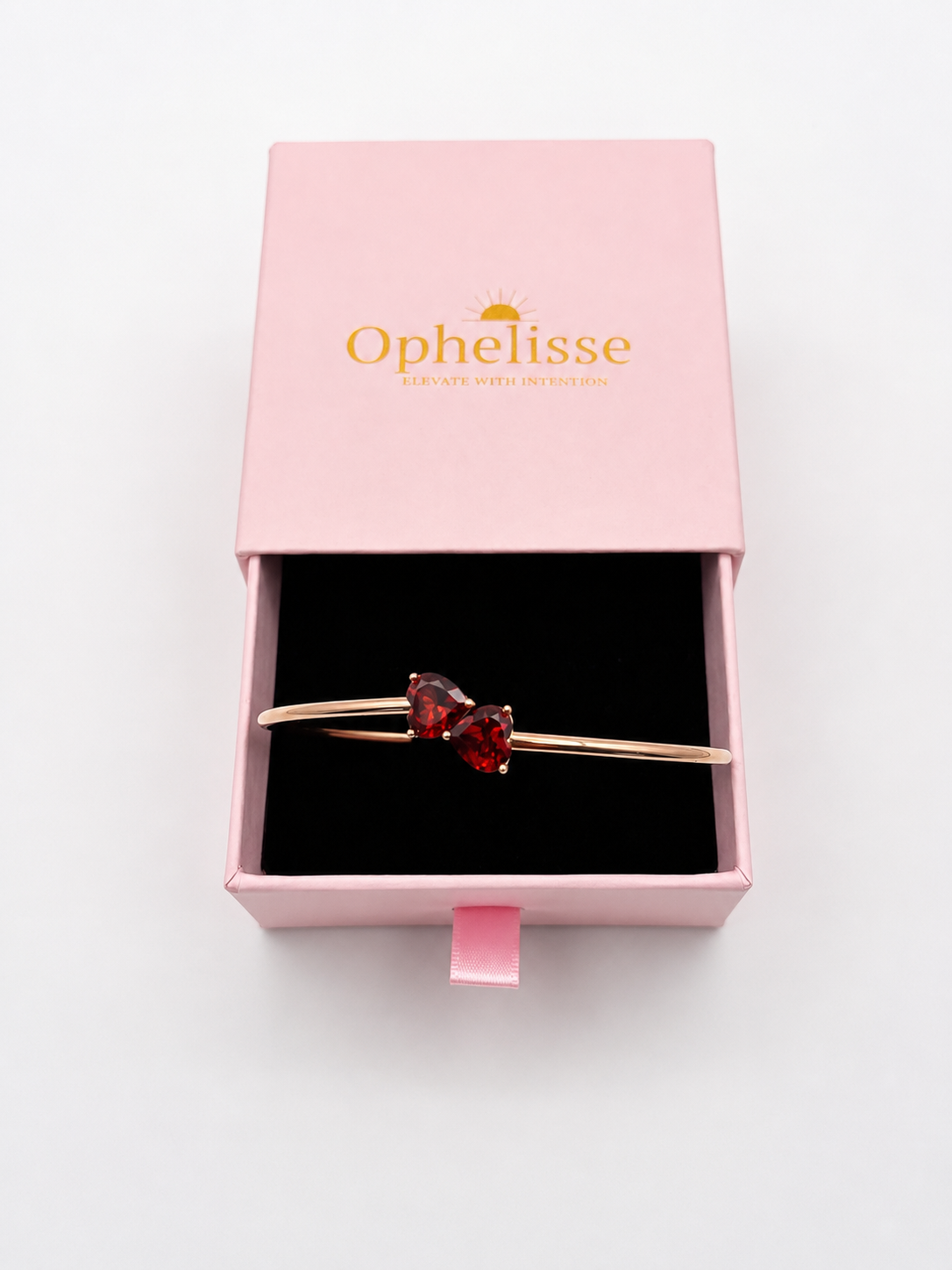 A solid 10k rose gold bangle bracelet with two red heart-shaped natural garnet gemstones, presented in an open pink Ophelisse gift box on a white background.