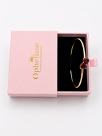 A solid 10k rose gold bangle bracelet with a double heart charm set with deep red natural garnet gemstones, presented in a pink Ophelisse gift box with a black interior, top view