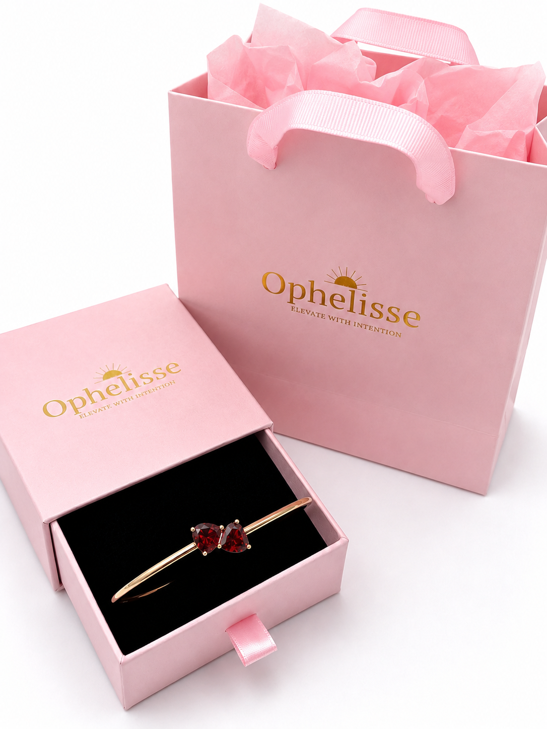 A solid 10k rose gold bangle bracelet with two natural red heart-shaped garnet gemstones displayed in an open pink Ophelisse jewelry box, with a matching pink shopping bag behind it on a white background.