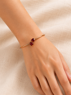 A close-up of a person's wrist wearing Ophelisse solid 10k rose gold bangle with two natural red heart-shaped stones, resting on a textured beige fabric background.