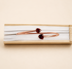 A solid 10k rose gold bangle bracelet with two small heart-shaped dark red natural garnet gemstones by Ophelisse, displayed on a warm beige background showing the top view of the bangle and natural garnet.