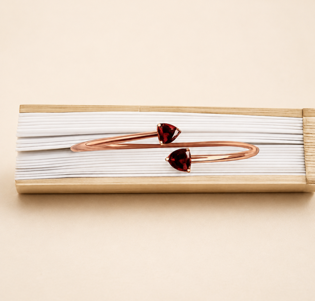 A solid 10k rose gold bangle bracelet with two small heart-shaped dark red natural garnet gemstones by Ophelisse, displayed on a warm beige background showing the top view of the bangle and natural garnet.