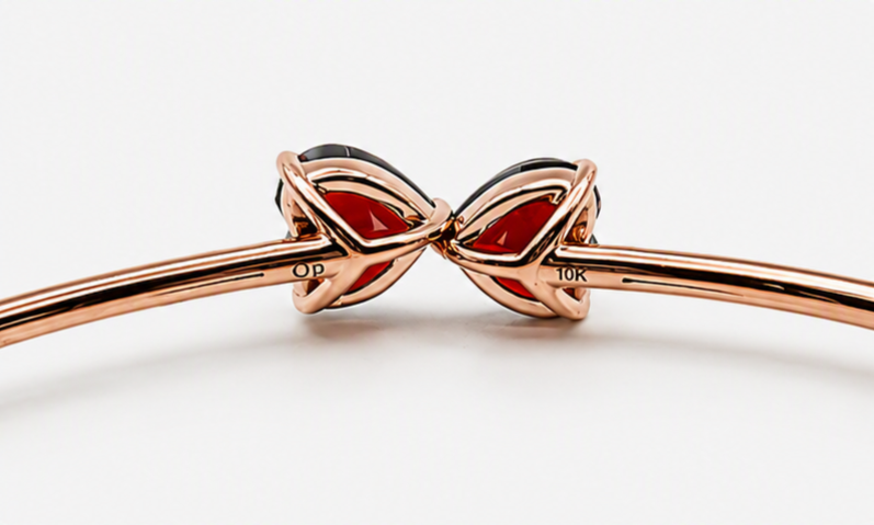 Close-up of a solid 10k rose gold bangle bracelet with a bow-shaped charm featuring natural red garnet, on a light gray background, showing Ophelisse trademark and 10k mark.