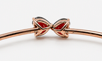 Close-up of a solid 10k rose gold bangle bracelet with a bow-shaped charm featuring natural red garnet, on a light gray background, showing Ophelisse trademark and 10k mark.
