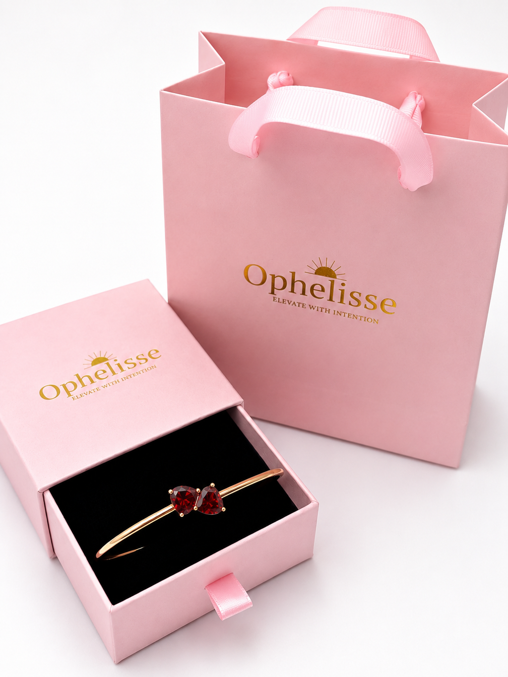 A solid 10k rose gold bangle bracelet with two red heart-shaped natural garnet gemstones, presented in an open pink Ophelisse jewelry box next to a pink branded shopping bag on a white background.
