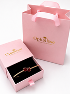 A solid 10k rose gold bangle bracelet with two red heart-shaped natural garnet gemstones, presented in an open pink Ophelisse jewelry box next to a pink branded shopping bag on a white background.