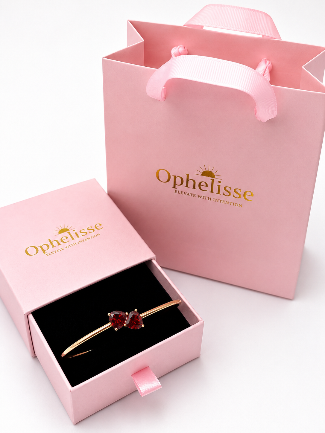 A solid 10k rose gold bangle bracelet with two red heart-shaped natural garnet gemstones, presented in an open pink Ophelisse jewelry box next to a pink branded shopping bag on a white background.