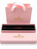 A solid 10k rose gold chain bracelet with natural red garnet heart charms is displayed in an open Ophelisse pink jewelry box, with a matching pink branded gift bag behind it on a white background.