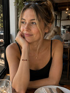 A woman sitting at a cafe table wearing Ophelisse's chain bracelet on her wrist, featuring solid 10k rose gold with two heart-shaped and two round-shaped natural red garnet gemstones.