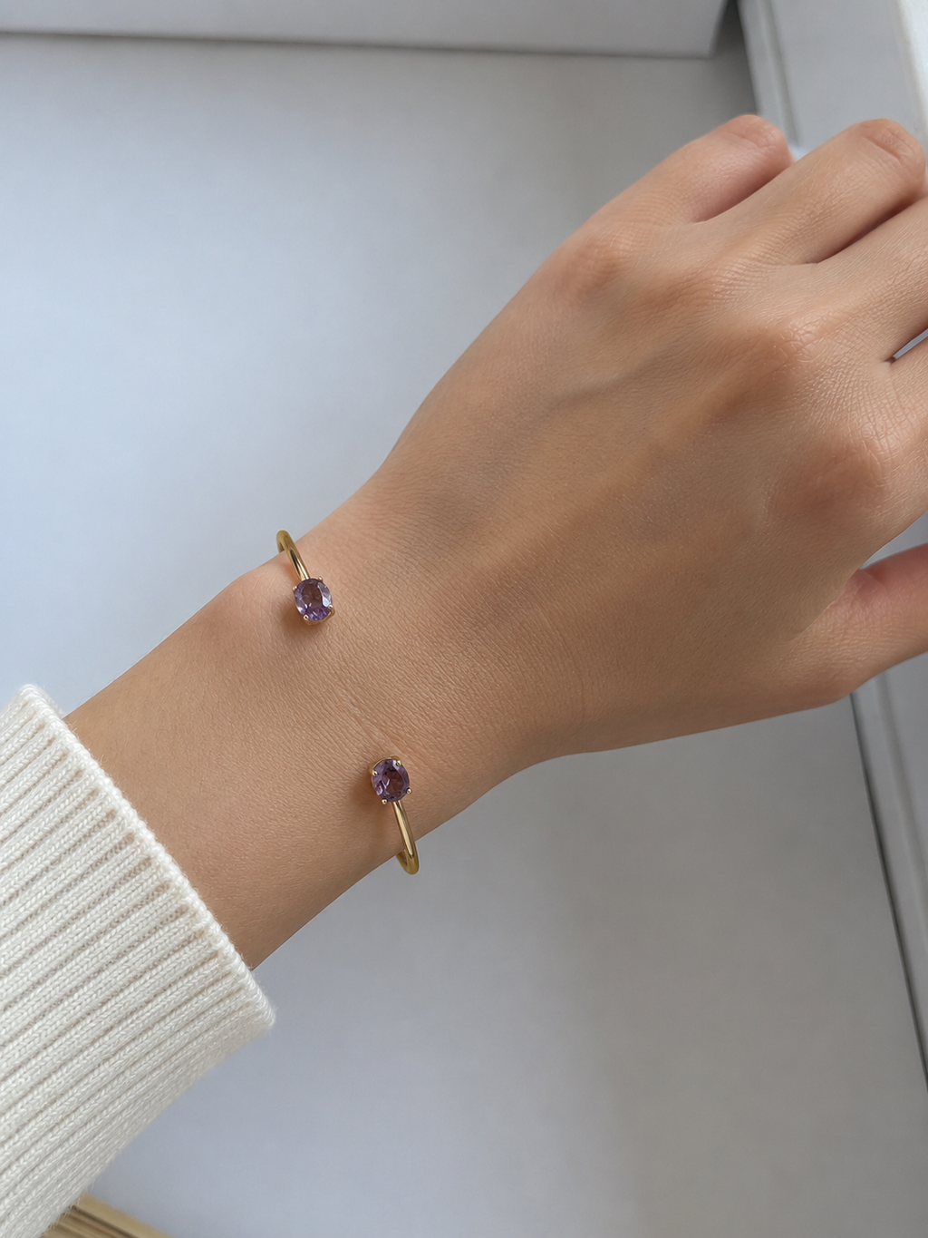 A close-up of a person's wrist wearing Ophelisse 10k solid yellow gold bangle with two oval natural amethyst gemstones, against a soft light gray background.