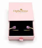 A solid 10k yellow gold cuff bracelet with natural oval purple amethyst gemstones, presented in an open pink Ophelisse jewelry box with a black interior, shown in top view.