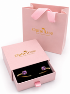 Ophelisse brand jewelry gift set featuring solid 10k yellow gold bangle with natural oval purple amethyst gemstones displayed in an open pink presentation box, with a matching pink shopping bag in the background.